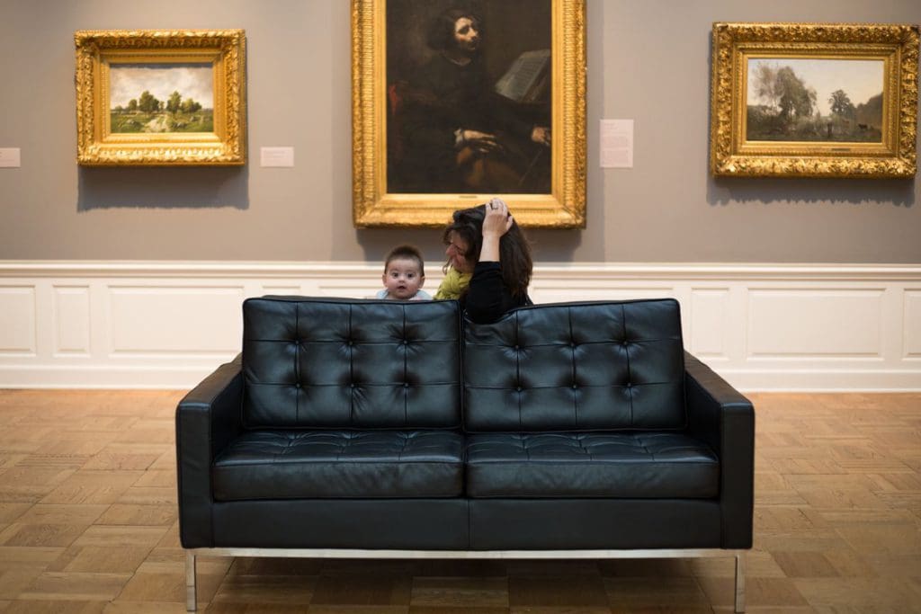 A mom and her infant sit on a couch at the Portland Art Museum, with three painting on the wall behind them.