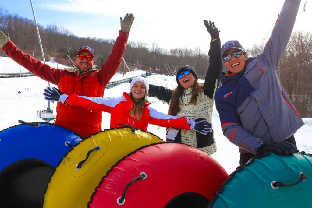 Four people, each standing behind their colorful tubes, throws their hands in the air in excitement on a winter day at Shawnee Mountain Ski Area.