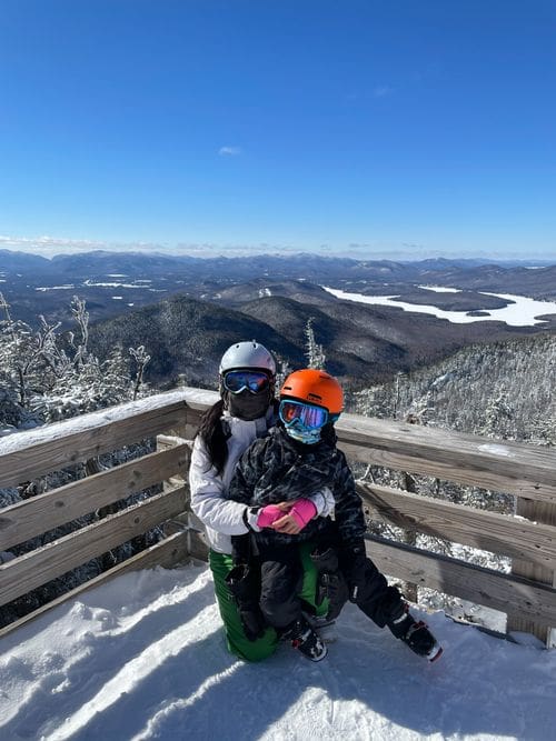 A mom and her young son, wearing full ski gear, stand together with a view of Whiteface Mountain behind them.