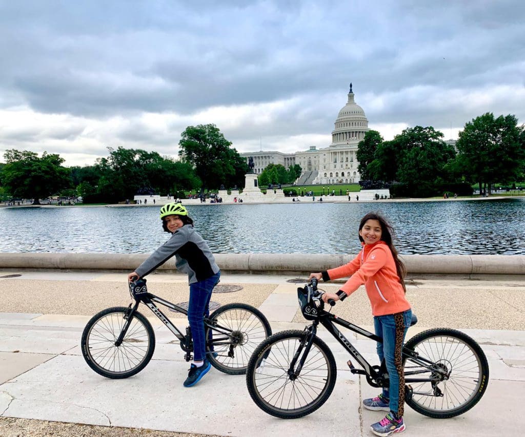 Two kids on bikes rest along the National Mall with monuments in the distance.