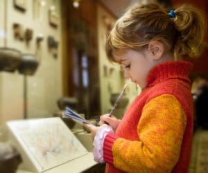 A young girl looks down at a museum exhibit, writing notes.