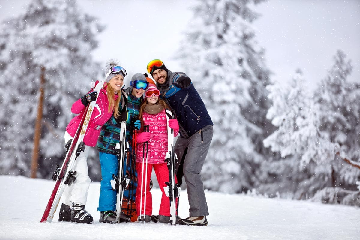 A family of four, all wearing colorful ski gear and skis, stands proudly on a winter landscape near Aspen, one of the best ski resorts in the United States for families.