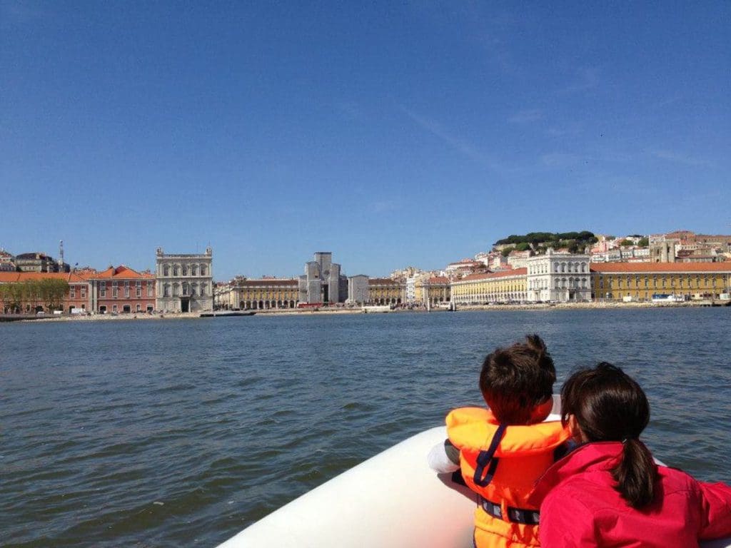 Two kids look over the bow of a ship toward a view of Lisbon from the water.