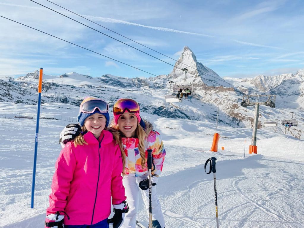 A young girl and her mom stand together, both wearing ski gear, on a sunny, winter day near the Matterhorn.
