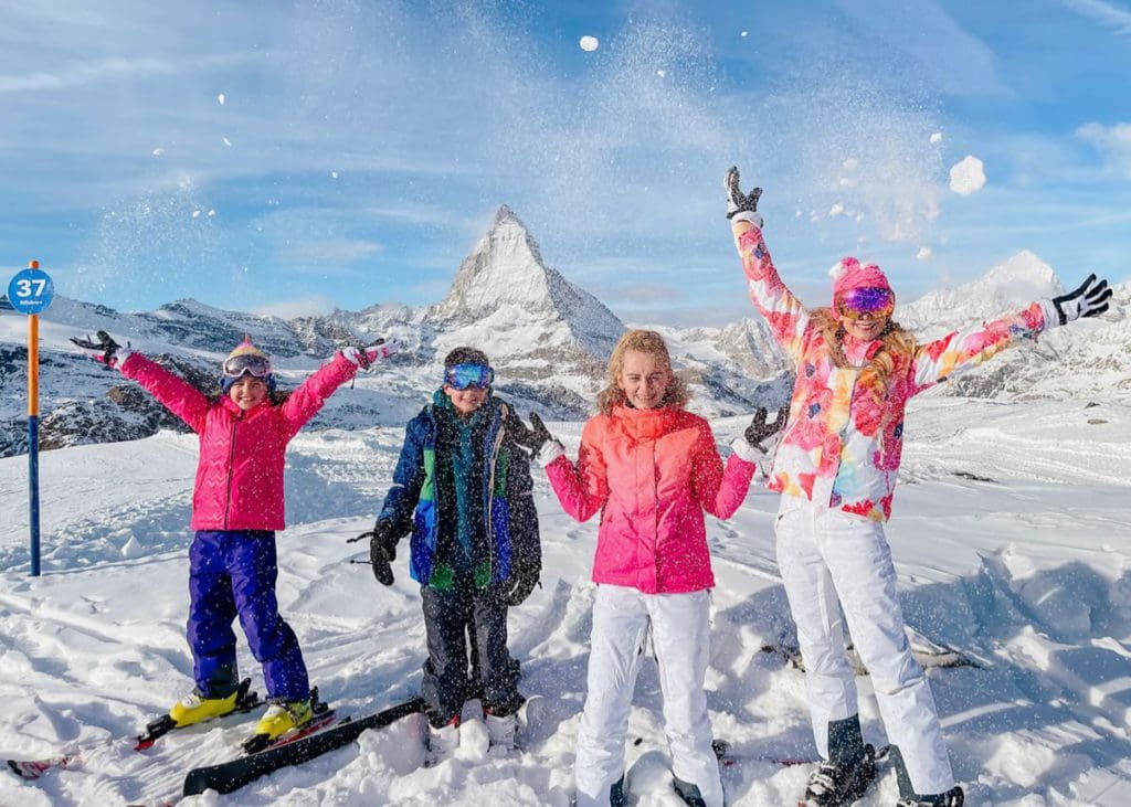 A mom and her three kids throw snow in the air, while skiing in Zermatt together at one of the best places to ski Europe with Kids.
