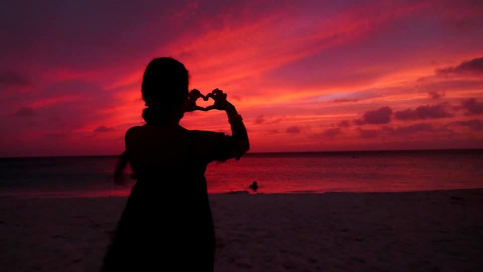 Someone stands on Eagle Beach making her hands into a heart at the ruby red sunset at one of the best beaches in Aruba for families.