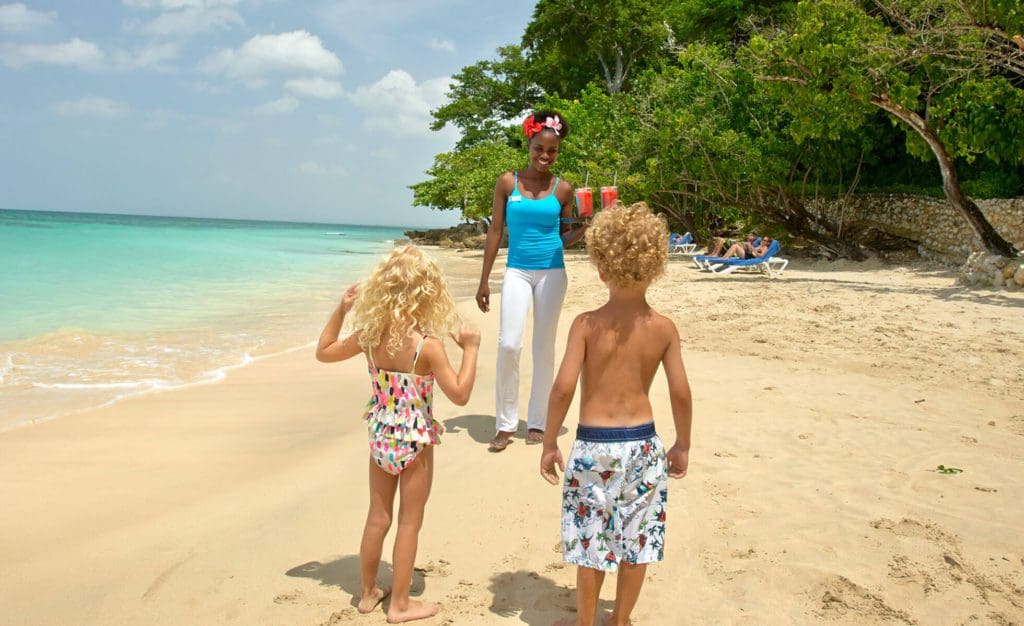 Two kids in swimwear walk toward a staff member at the Bluefields Bay Villas, holding smoothies for them on the beach.