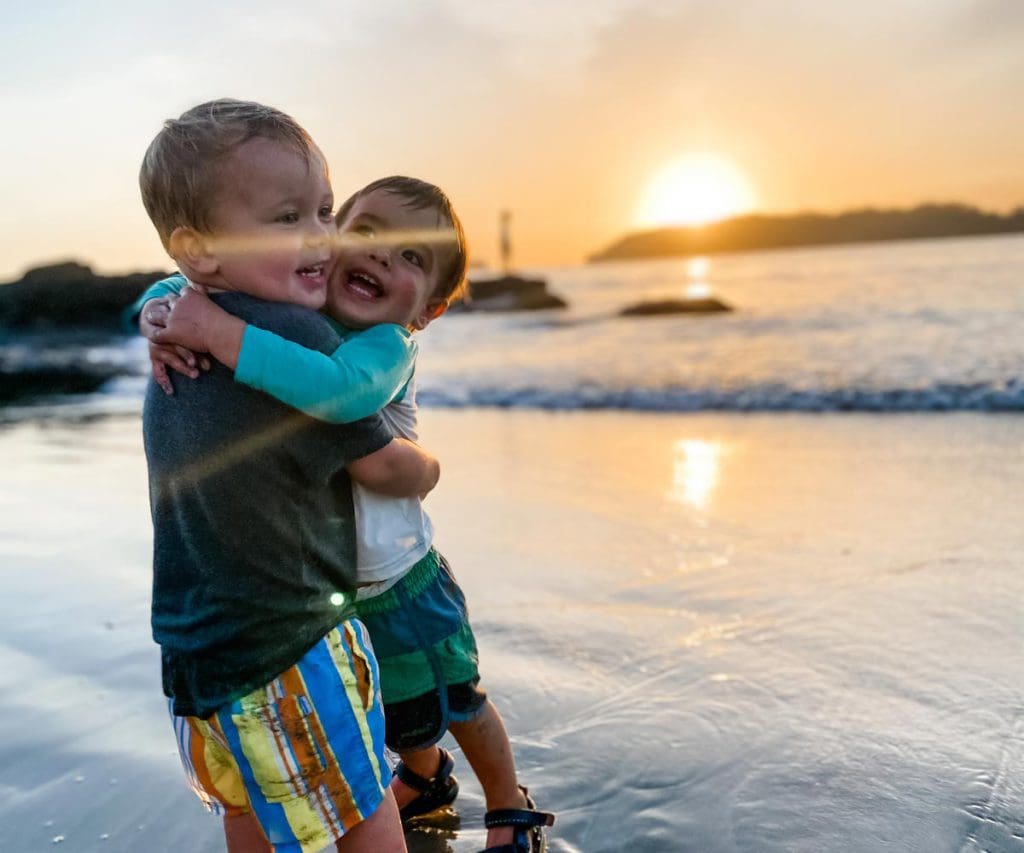 Two toddler boys embrace on the beach at sunset.