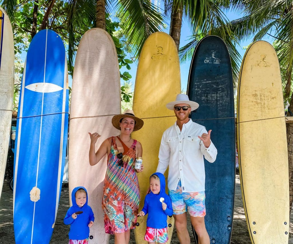 Two parents and two toddler boys stand in front of five colorful surfboards.