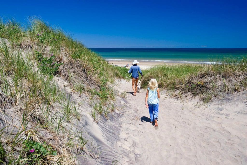 A young child chases after her dad along a sandy path between beach grass in Block Island, one of the best cool-weather destinations in the United States for families.