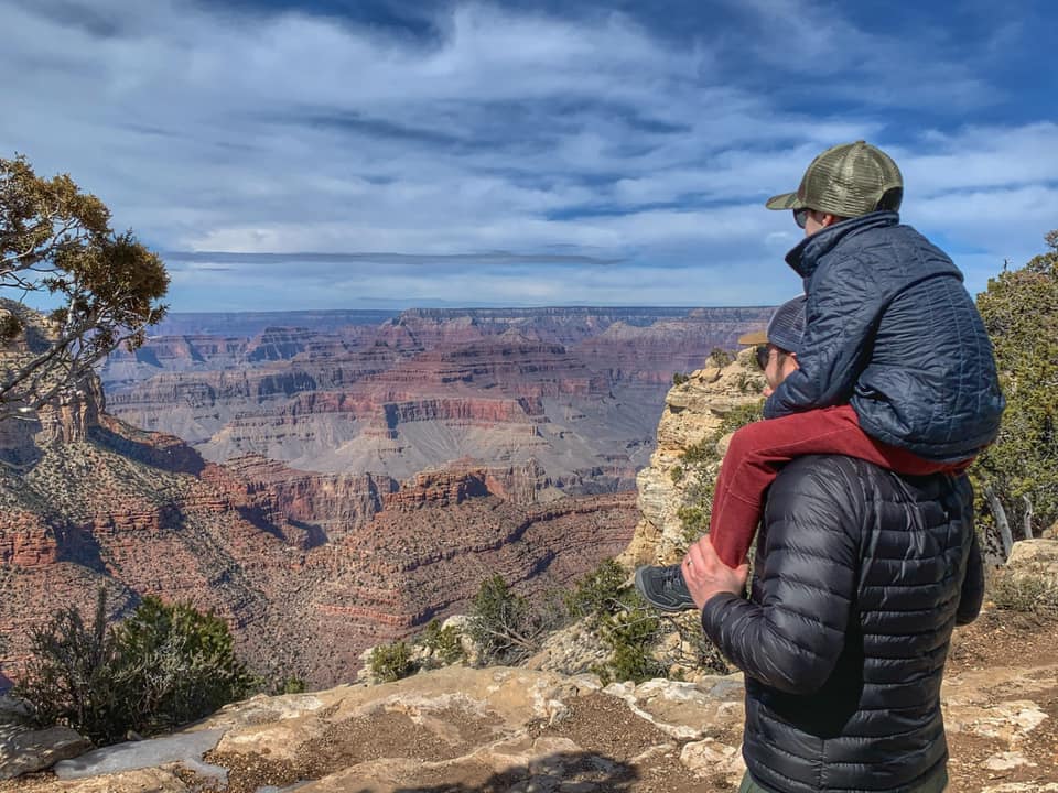 A dad holds his young son on his shoulders as they look out onto the Grand Canyon.