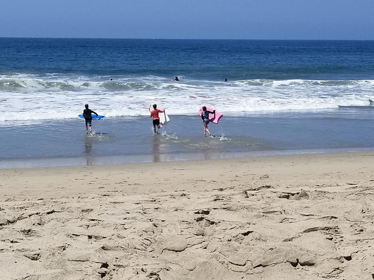 Three kids race to the ocean with boogie boards in their hands on a beach near LA, one of the best US cities for a Memorial Day Weekend with kids.