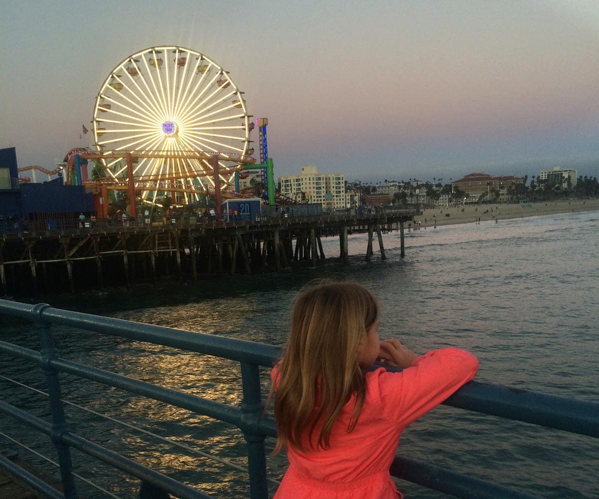 A young girl leans over a railing while looking at the lit up Ferris wheel at the Santa Monica Pier.