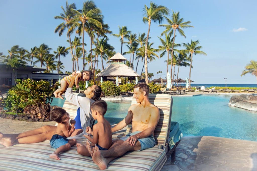 A family of five sits together, making memories, on pool loungers near the pool at Fairmont Orchid, Hawaii on a sunny day.