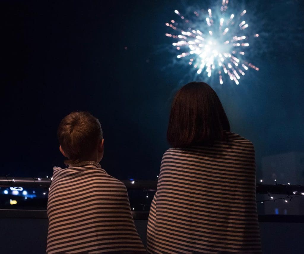 Two kids, wrapped in blankets, look up at Fourth of July fireworks on a family trip.