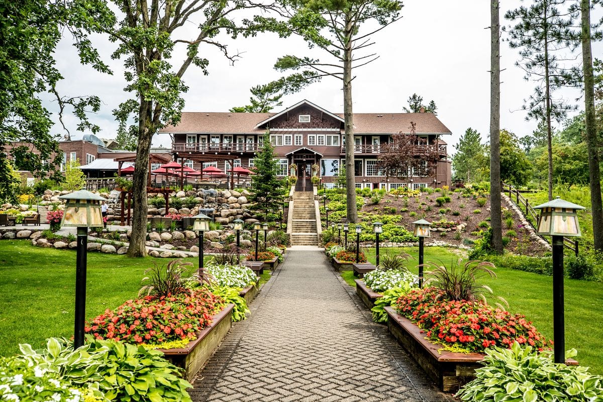 The grand exterior entrance to Grand View Lodge Spa & Golf Resort, featuring a long walk-up, flanked by flower beds on each side, with the lake in the distance.