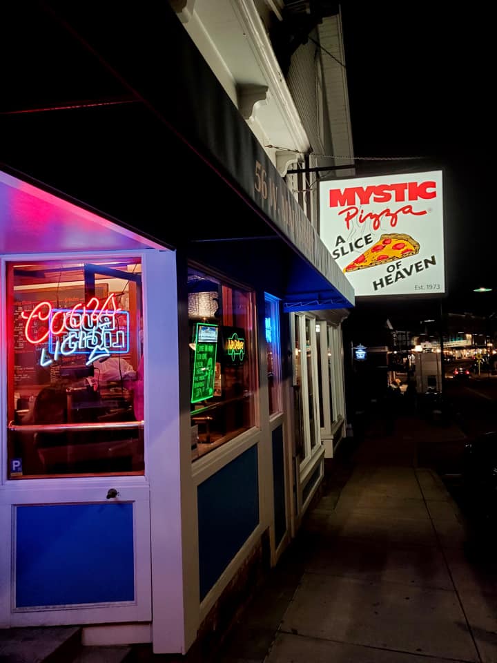 A view down the building side of Mystic Pizza, with the neon sign lit up at night.
