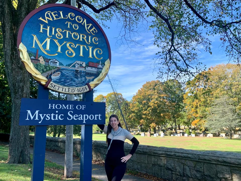 A teenage girl stands next to the welcome sign for Mystic on a sunny day.