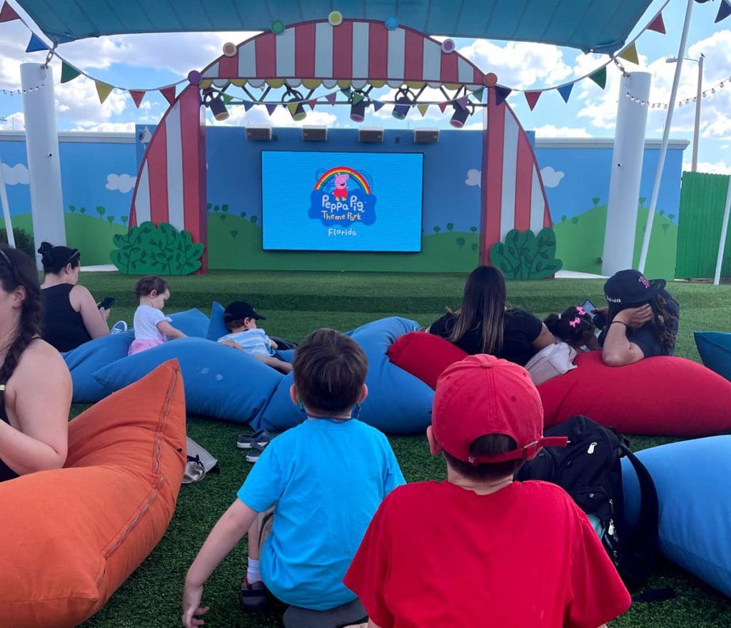 Several families sit on the ground at an outdoor move theatre featuring Peppe Pig films.