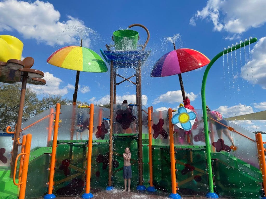 A young boy stands under the falling water at a brightly colored splash pad attraction that dumps water from overhead.