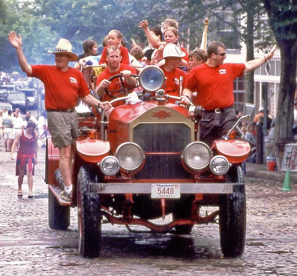Several people dressed in red wave from a red, old-fashioned car during a Fourth of July parade in Nantucket, one of the best Fourth of July destinations for a family trip.