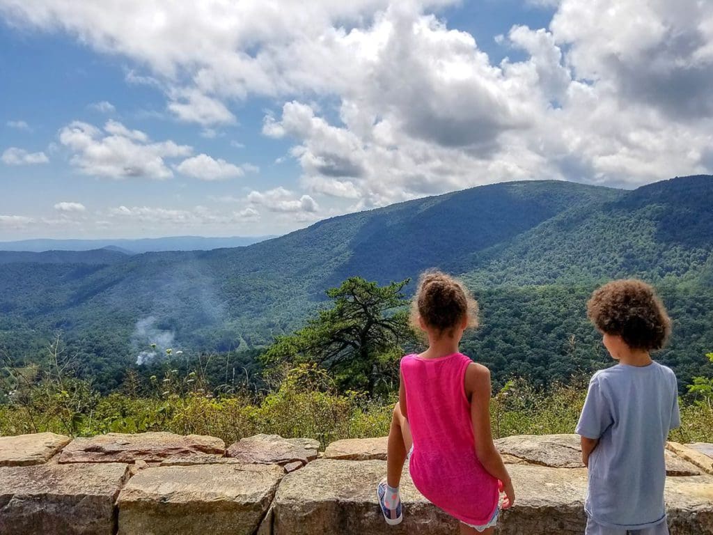 Two kids stand along a rock fence in Shenandoah National Park, looking over at a few of the mountains.