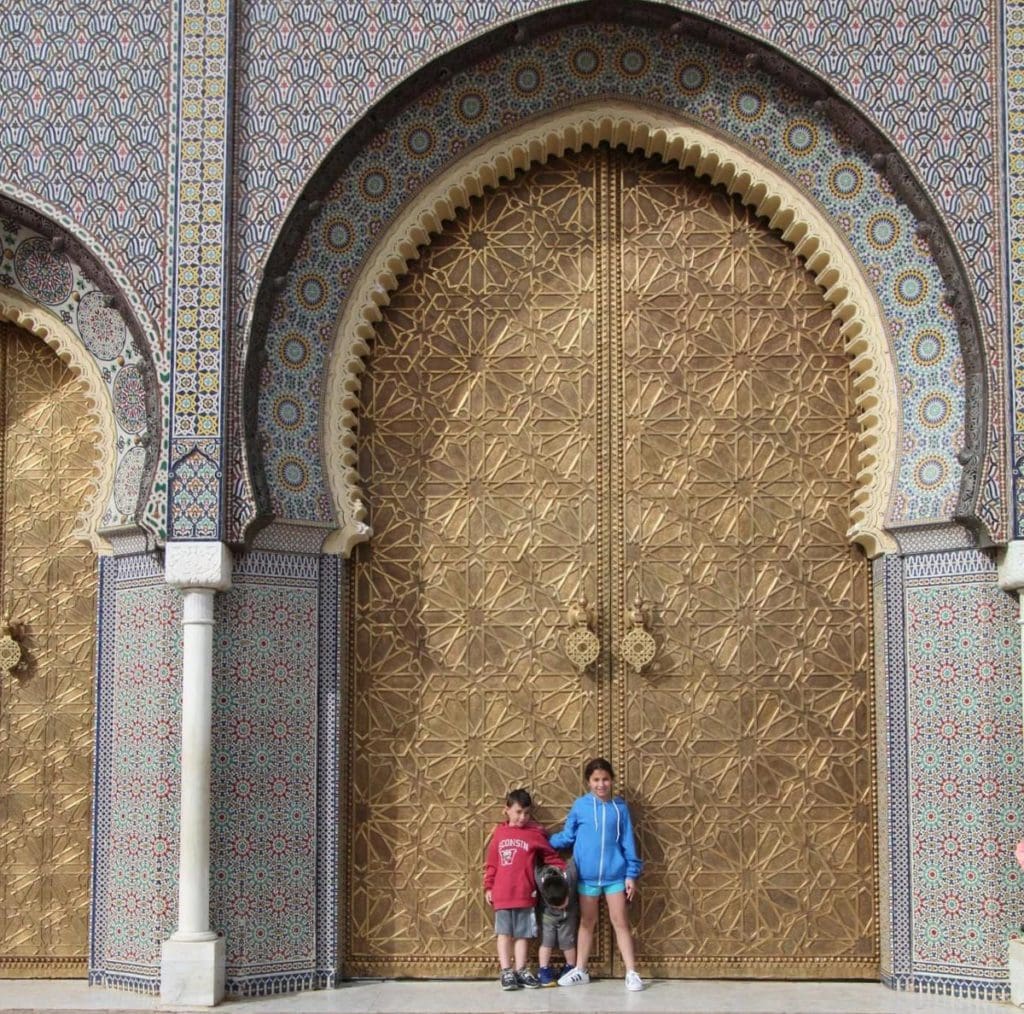 Two kids stand in front of a grand door at a Moroccan mosque.