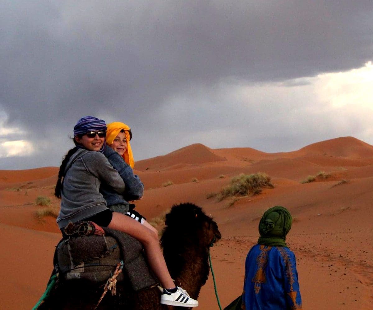 A guide leads a camel, atop of which are a mom and child, in the desert of Morocco.