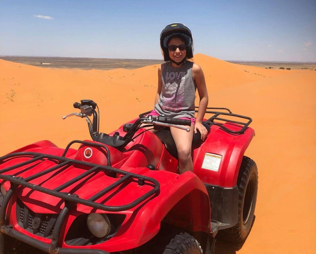 A young girl wearing a helmet sits atop a red four-wheeler in the Saharan Desert.