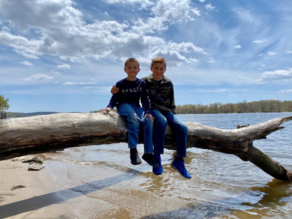 Two kids sit together on an overturned tree, reaching into the waters of a river at Gillette Castle State Park in Great Barrington, one of the Best Cute Towns To Visit With Kids Near NYC.