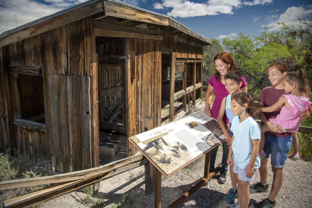 A mom and two kids look on at an informational display near a cabin exhibit at Springs Preserve.