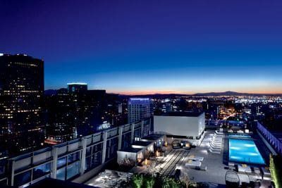 The rooftop pool and surrounding pool deck at The Ritz-Carlton, Los Angeles, with a view of the LA skyline at night.