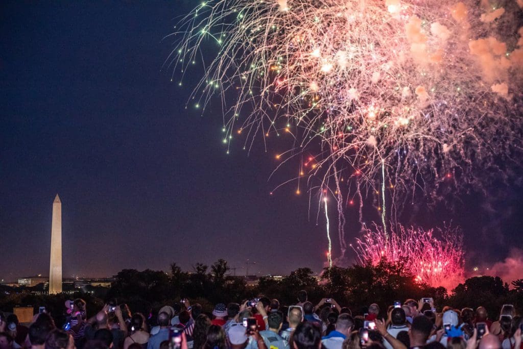 A huge crowd of people enjoy fire works, while the Washington Monument can be seen in the distance, one of the best Fourth of July destinations for a family trip.