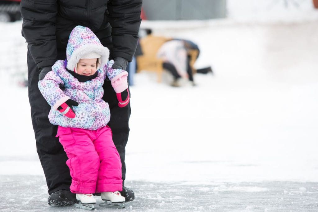 A young toddler learns to sake, while an adult holds onto their arms and skates behind her.