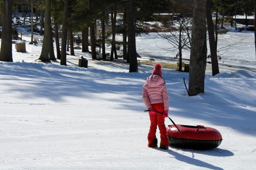 A young girl wearing a pink coat walks along a winter scene, pulling a red snow tube behind her.
