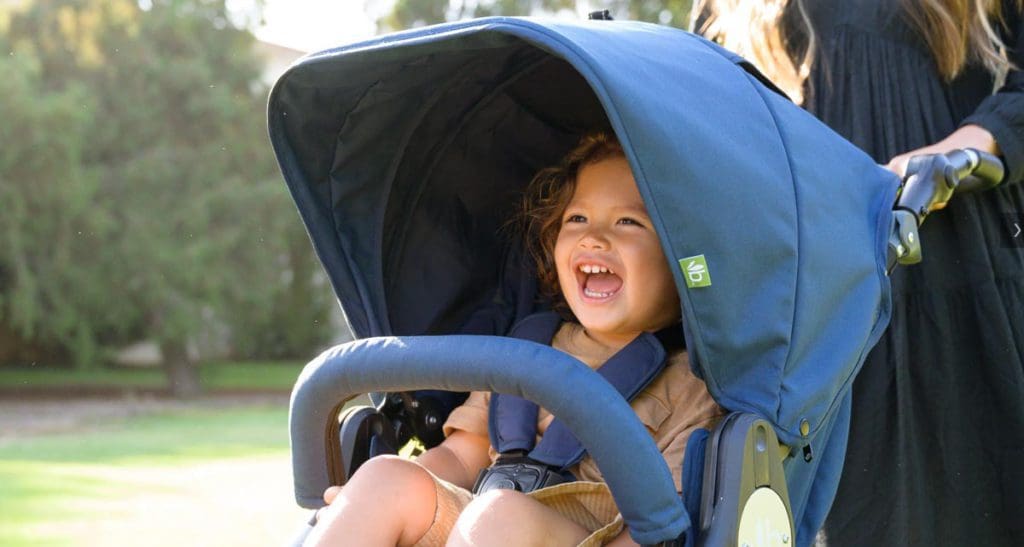 A close up of a laughing toddler in a Bumbleride Indie All-Terrain Stroller, with moms hands show pushing the stroller outside.