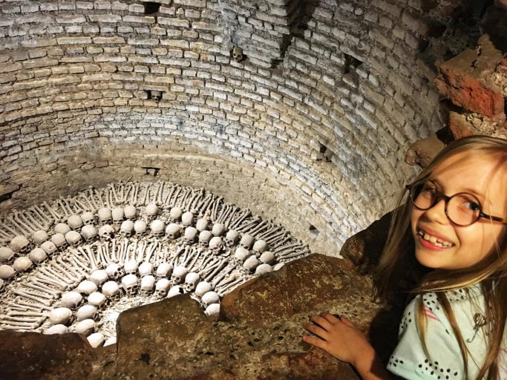 A young girl stands above a catacomb area, holding several ancient skulls.