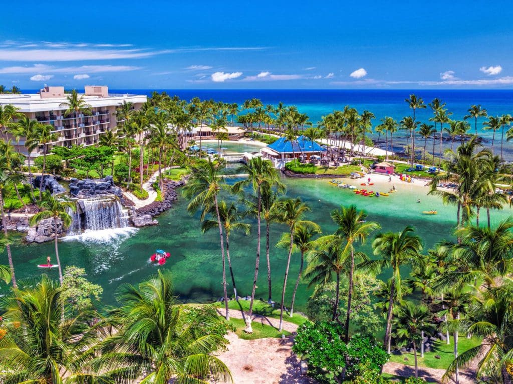 An aerial view of Hilton Waikoloa Village, featuring a large swimming area, ocean view, and resort buildings amongst lush greens.