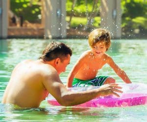 A father and son play together in a pool, using a large pink flotation device.