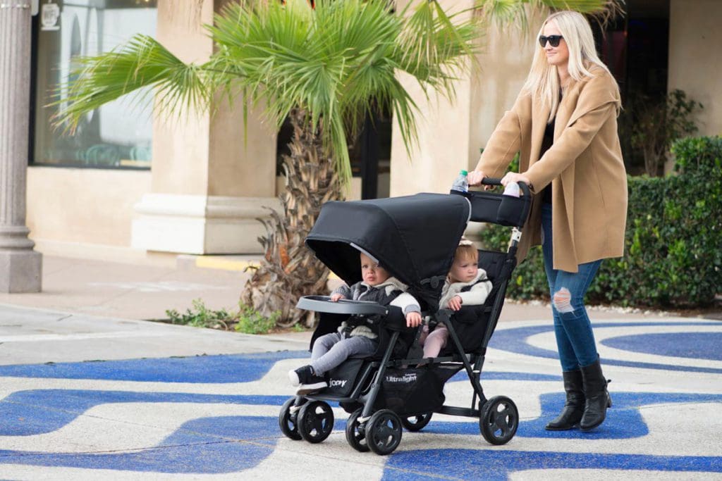 A mom pushes her two kids in the Joovy Caboose Too Ultralight Stand-On Stroller on a tropical city street.