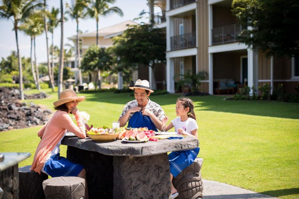 A family of three sits together and has lunch on the green lawn of the Kings’ Land, a Hilton Grand Vacations Club.