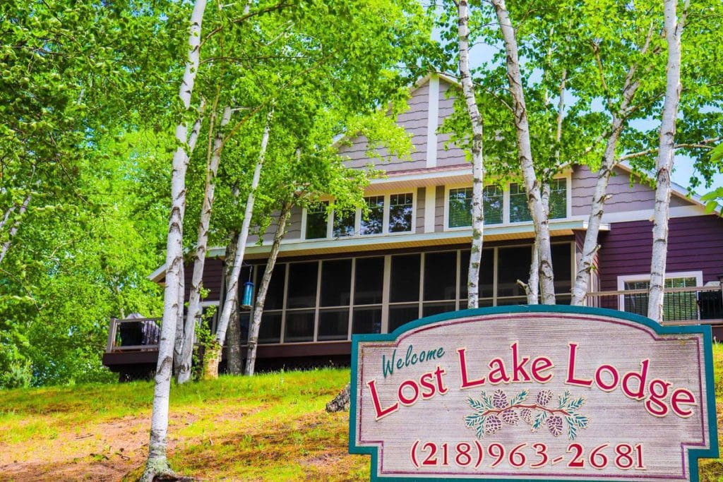 A sign reading Lost Lake Lodge sits in front of the welcome building, surrounded by full, green trees.