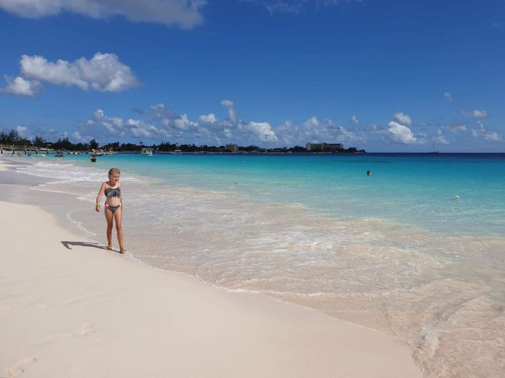 A young girl wearing a swim suit walks along the shore of the ocean in Barbados, with a large shoreline stretching out behind her.