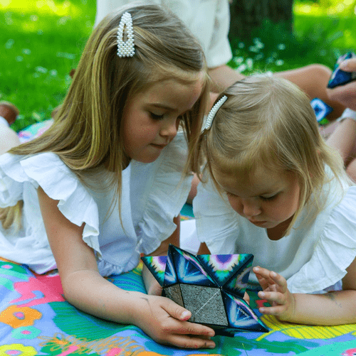 Two young girls play with a SHASHIBO Shape-Shifting Box together on a picnic blanket.