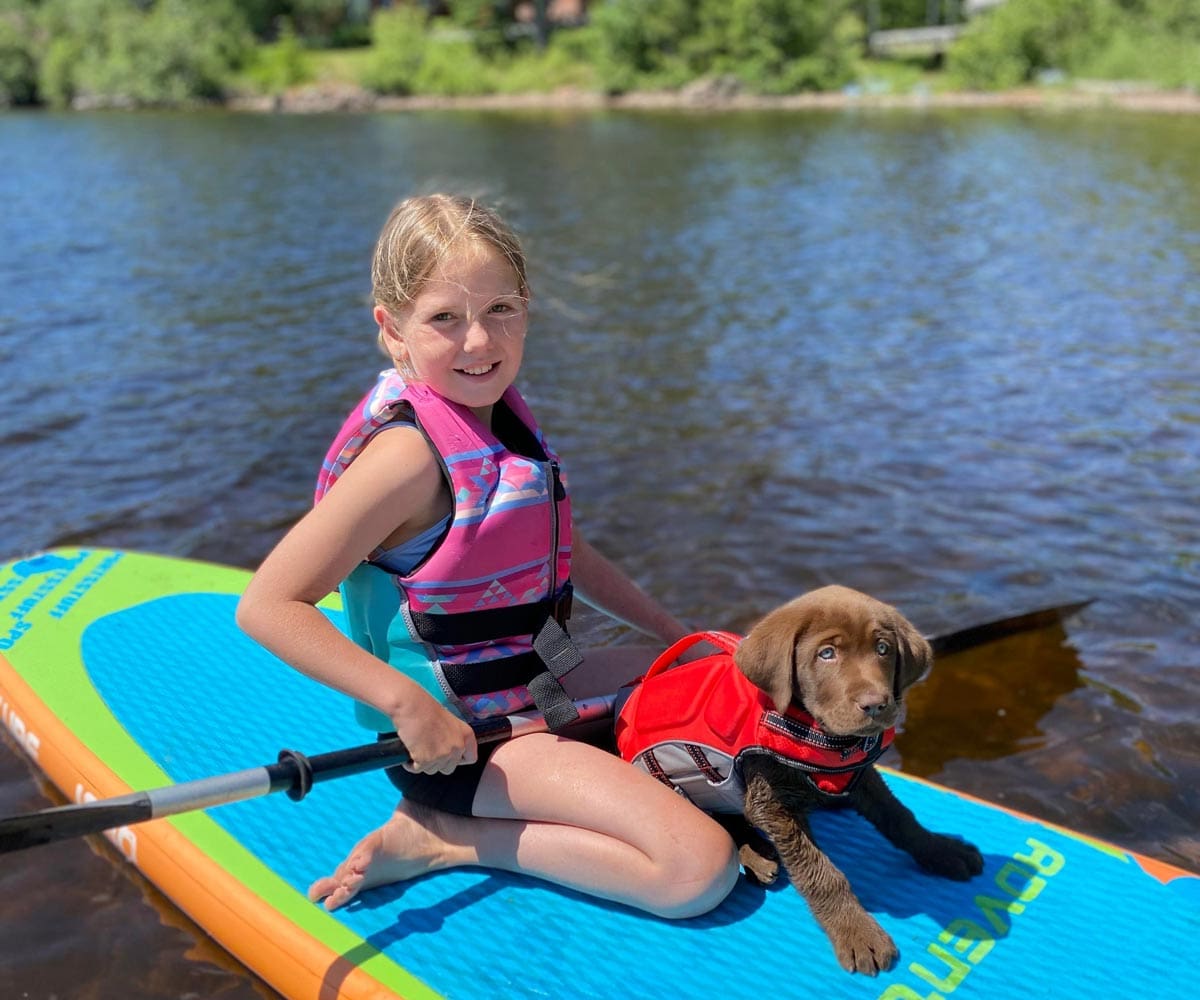 A young girl and her small Chocolate Lab puppy sit on a paddleboard on Devil Track Lake, while staying at Skyport Lodge.