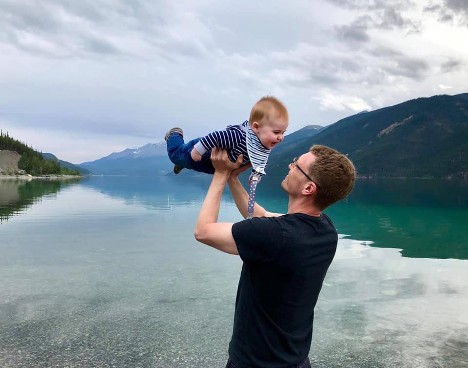 A dad raises his young baby overhead with Muncho Lake in British Colombia in the distance, , one of the best vacation destinations for families In Canada.