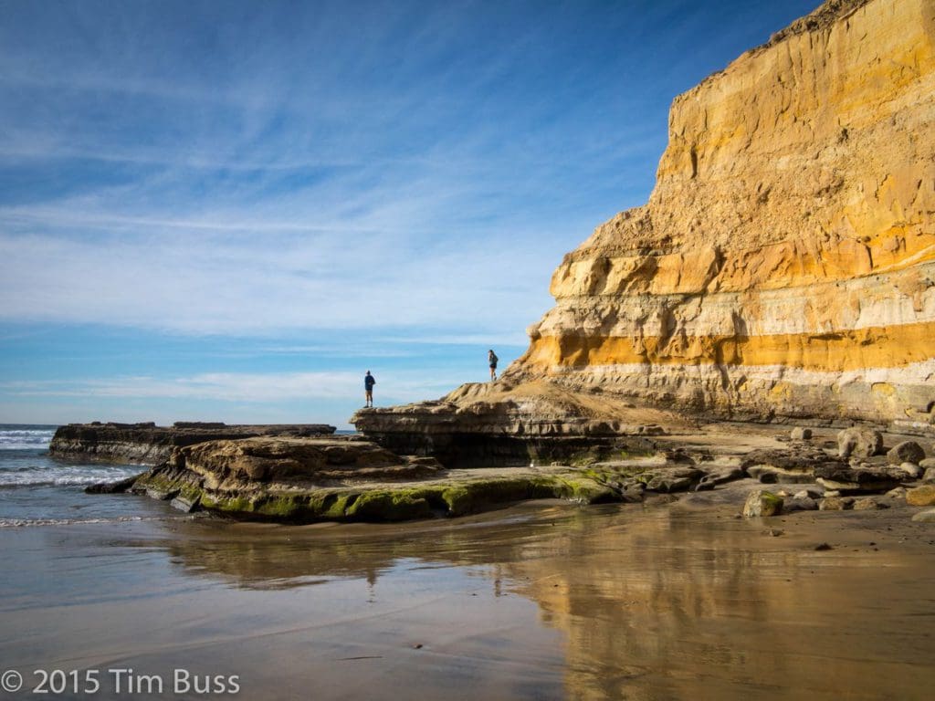 Two people explore large rocks stretching out into the ocean, with a cliffside behind them at Torrey Pines State Beach.