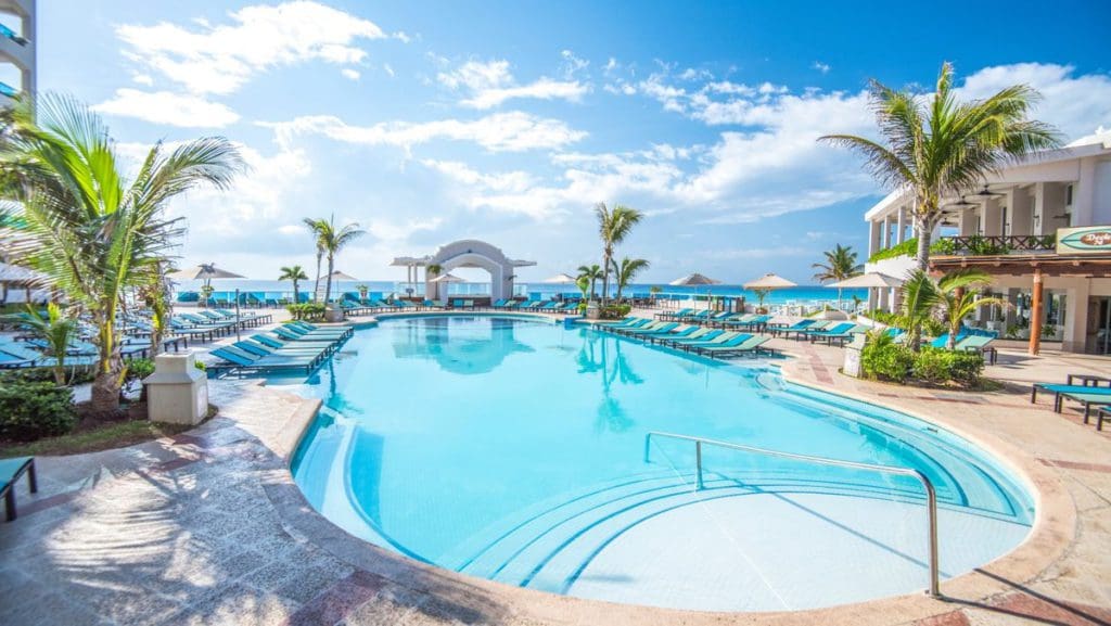 The pool and surrounding pool deck at Wyndham Alltra Cancun, featuring a beautiful arch to the ocean and swaying palm trees.