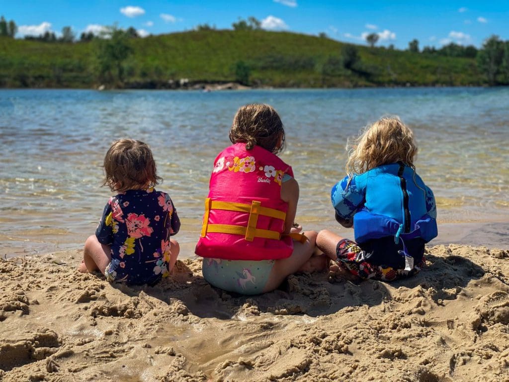 Three kids in swim suits and life jackets sit at the edge of Lake Wazee in Wisconsin.