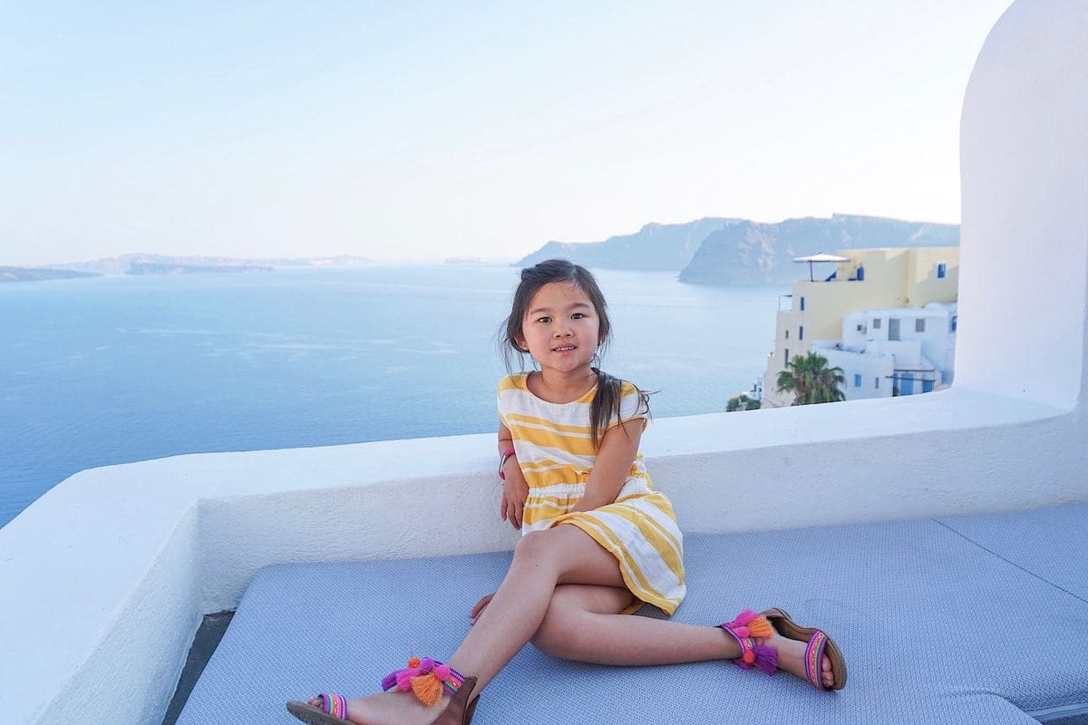 A young girl sits on a terrace in Greece with buildings and the ocean in the distnace.